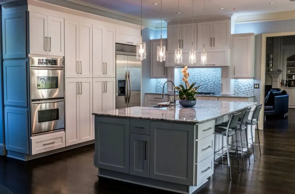 Kitchen by MG Builders with white shaker cabinets, double wall ovens, a gray island with a marble-look top and glass pendants